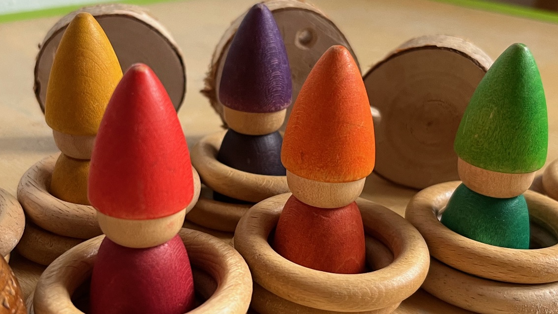 fünf verschiedenfarbige Figuren mit bunten Zipfelmützen aus Holz, die jeweils in einem Holzring stehen